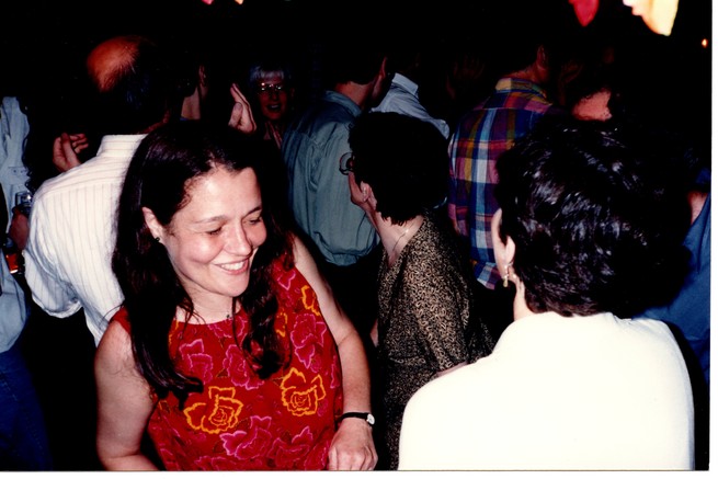 A dark and crowded dance floor; in the foreground, a dancing woman in a red dress smiles with her eyes closed, next to a woman in a white top who is not facing the camera.