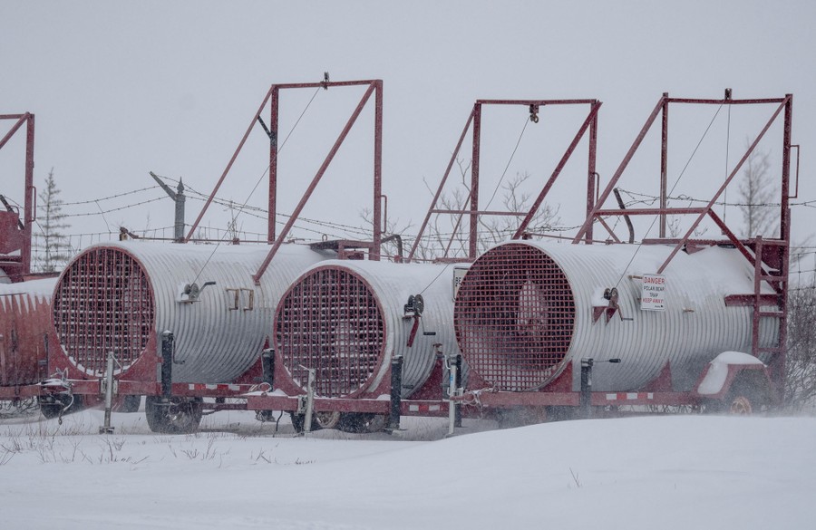 Several large cylinders mounted on trailers are seen in a parking area. The devices are cages, with rigging to open a front gate.
