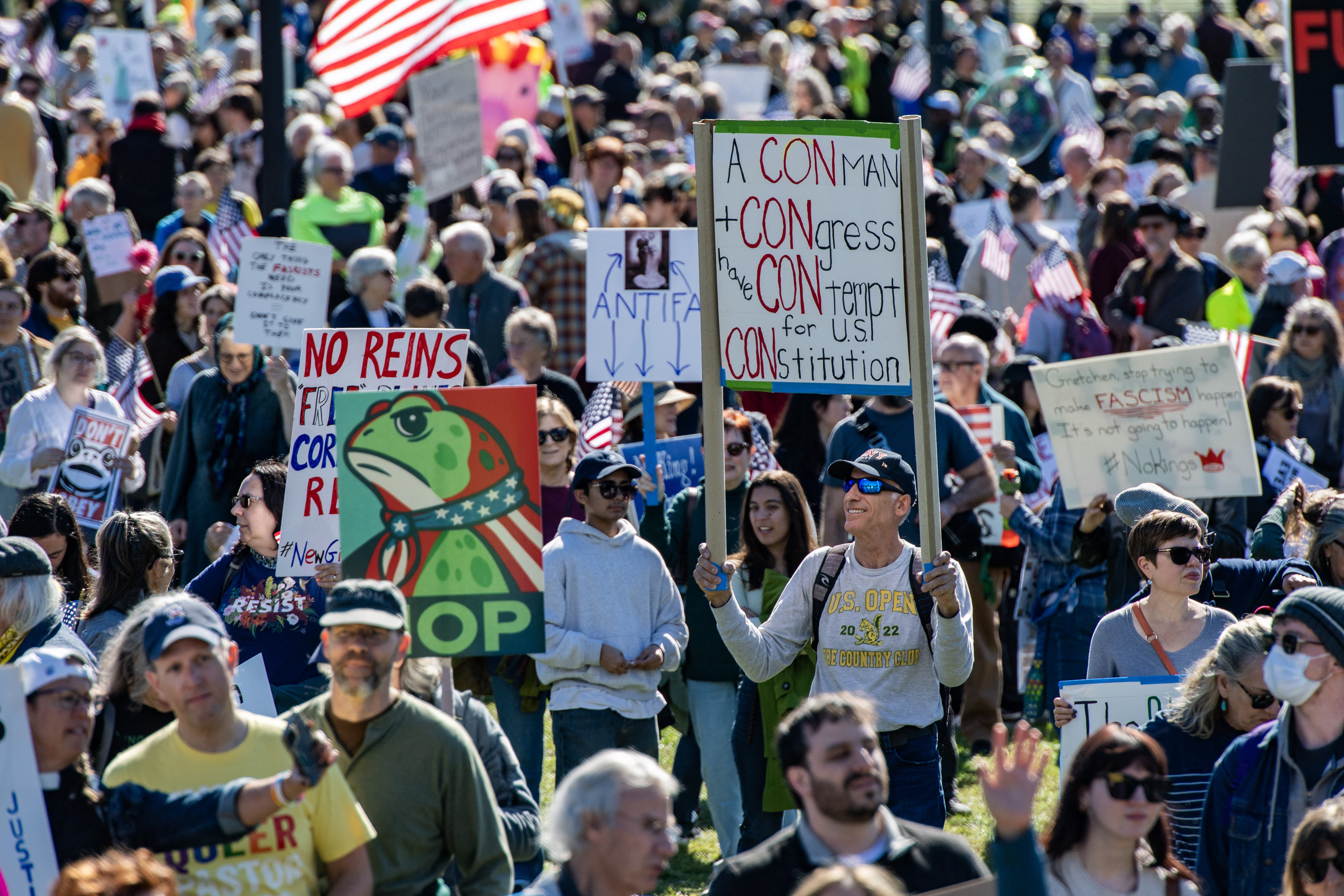 A large group of protesters gather in a park in Boston, holding signs, including one featuring a stylized cartoon frog and the word