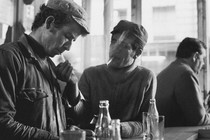 A black and white image of two men in a bar smoking. One man offers the other a light.