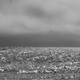 A black-and-white photograph of ocean waves below a stormy sky
