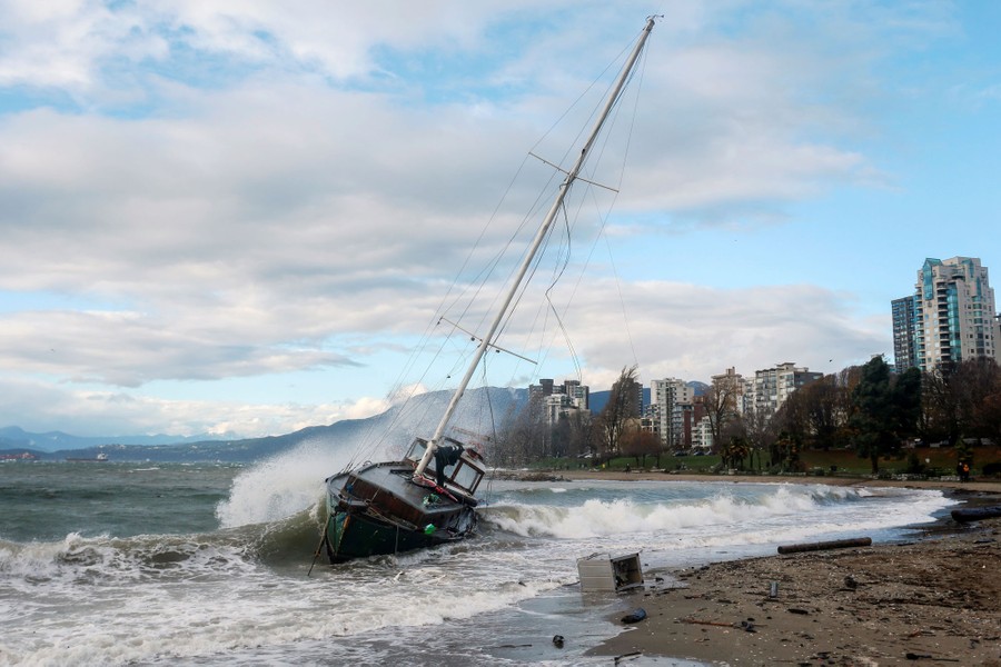 A sailboat rests on its side along a beach, bashed by waves.
