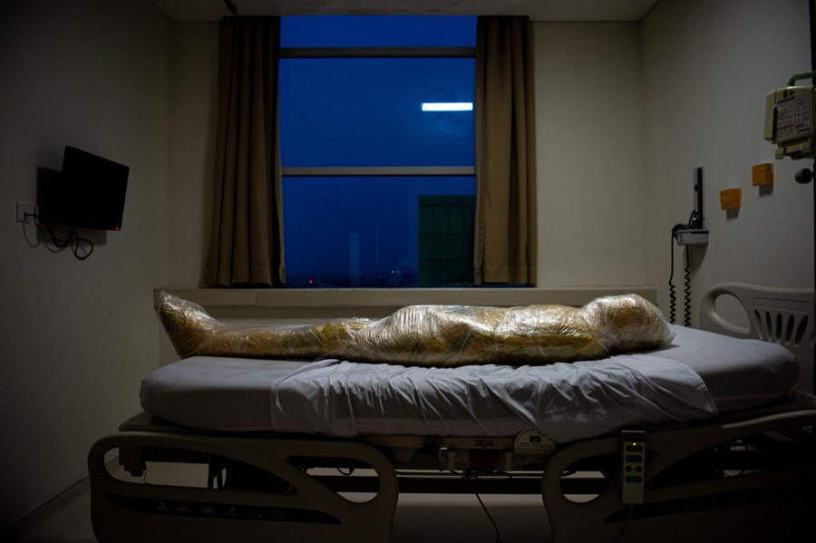 A body, wrapped in plastic, lies in a hospital bed.