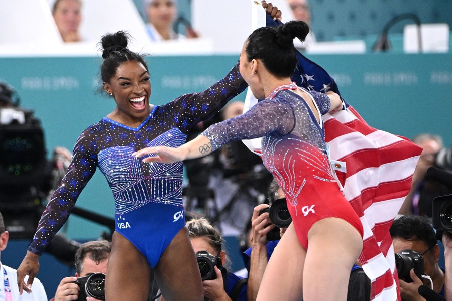 Two gymnasts celebrate, holding up an American flag, after winning Olympic medals.