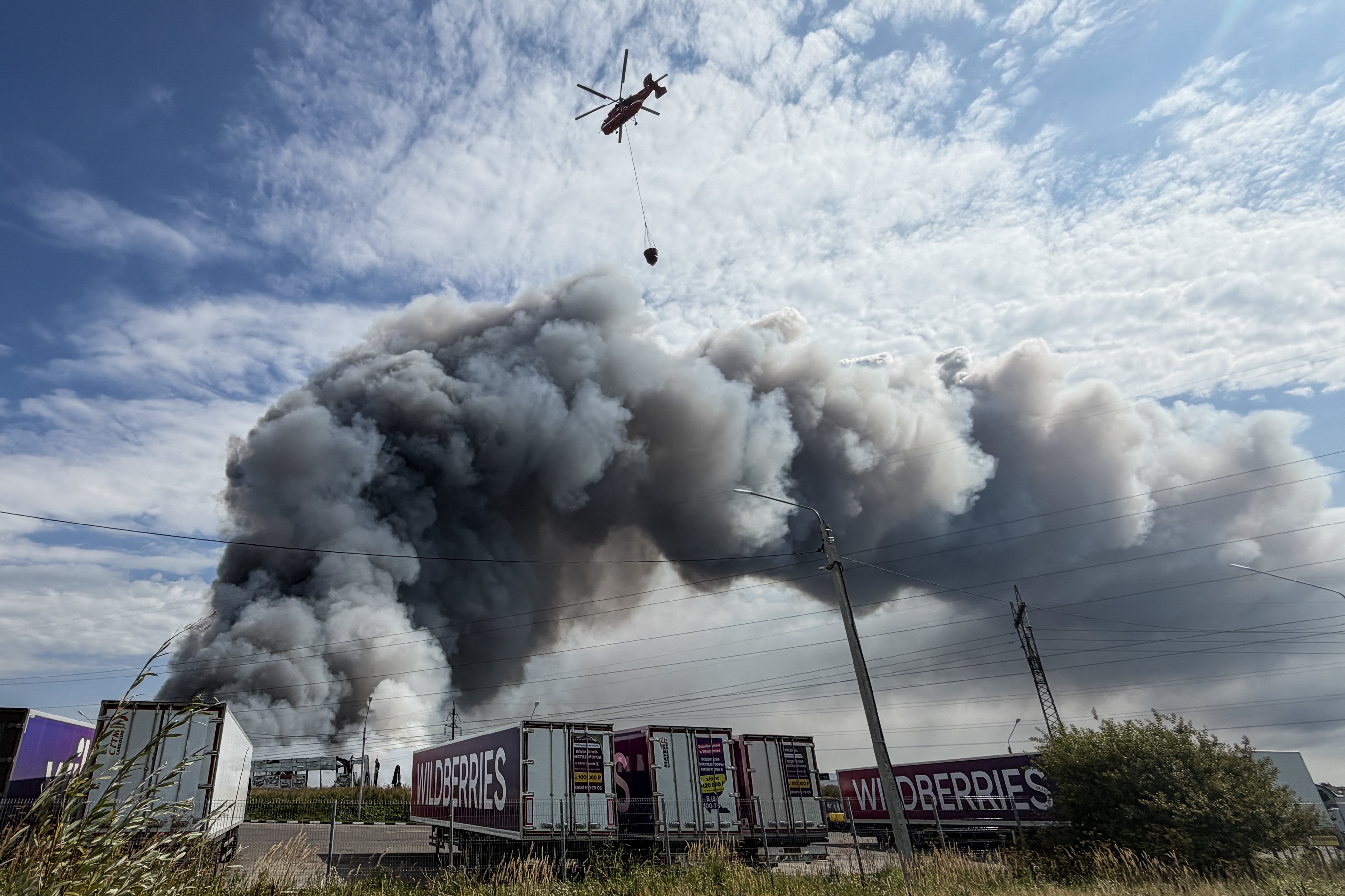 A firefighting helicopter lifts a bucket of water above a burning warehouse.