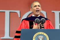 Barack Obama stands at a microphone wearing a graduation gown.