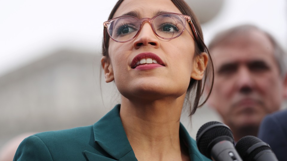 Alexandria Ocasio-Cortez stands at a bank of microphones in a green jacket.