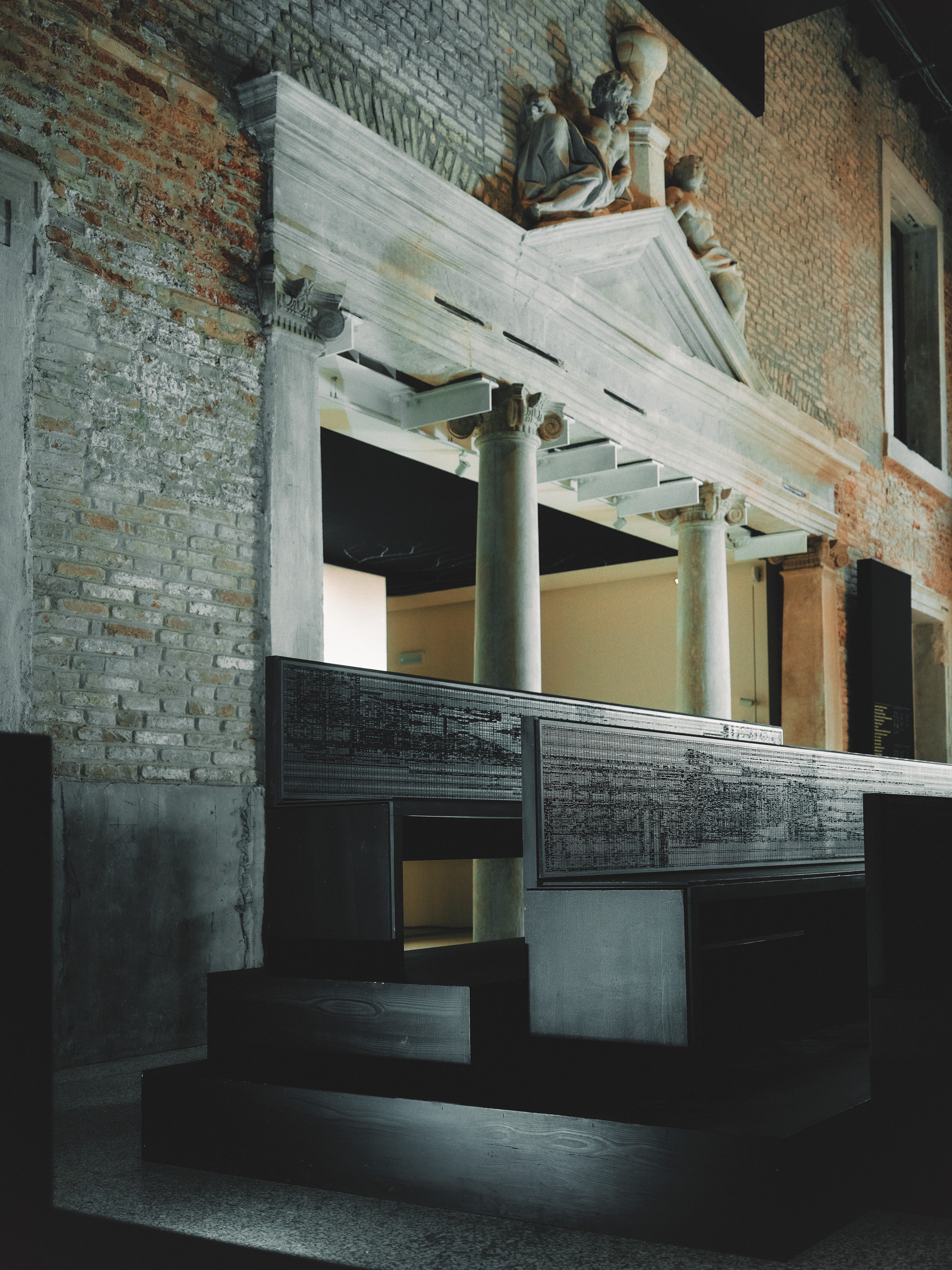 photo of installation with dark stair-stepped wooden benches in large brick hall with marble pediment and sculptures over wide doorway with marble columns