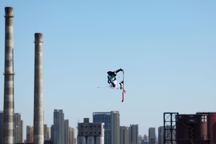 A skier is seen in midair, with tall buildings and smokestacks in the background.