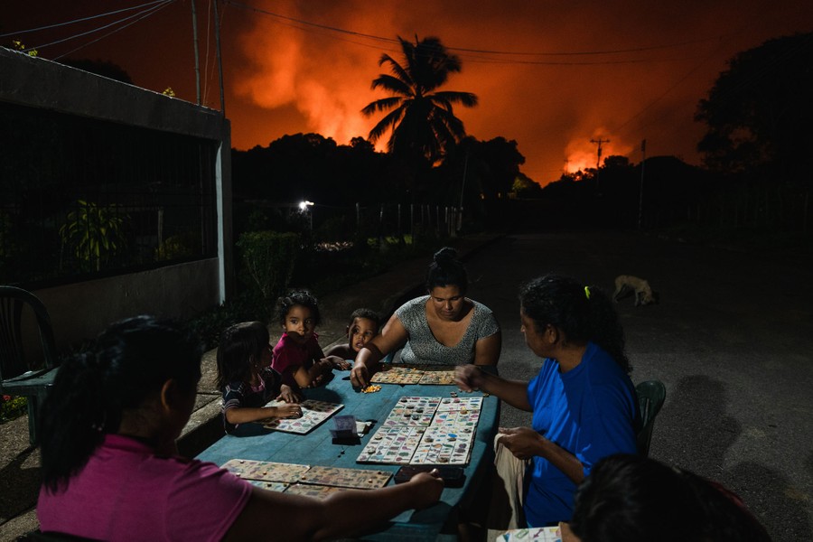 A family plays a board game at a table outside at night, the sky behind them partially lit by burning gas flares.