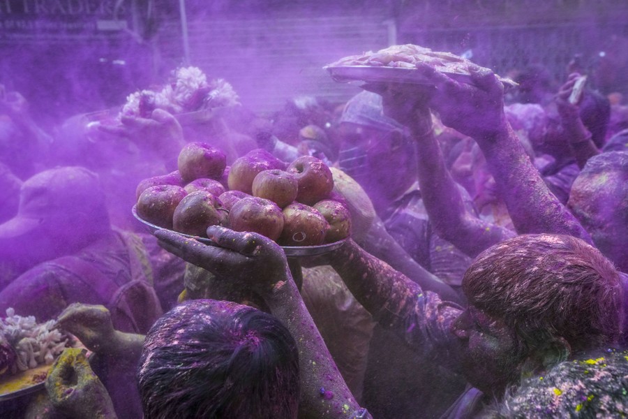 People covered in colored powder walk in a crowd; some carry offerings on plates.