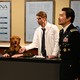 Still from 'Saturday Night Live' featuring SImu Liu in a military uniform, Cecily Strong dressed like a bureaucrat, and Mikey Day dressed like a researcher, all of them surrounding a dog with human hands wearing camouflage