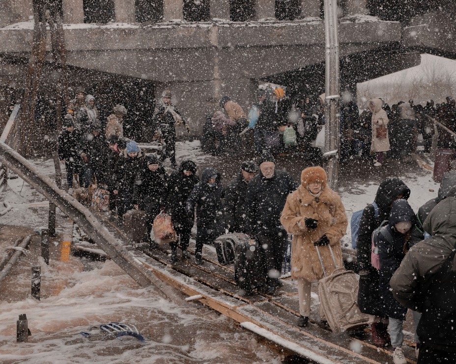 Snow falls on people trying to cross the detritus of a  bombed out bridge