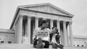 A black-and-white photograph of a woman and child sitting on the steps of the Supreme Court, holding up a newspaper with the front page headline, 'HIGH COURT BANS SEGREGATION IN PUBLIC SCHOOLS'
