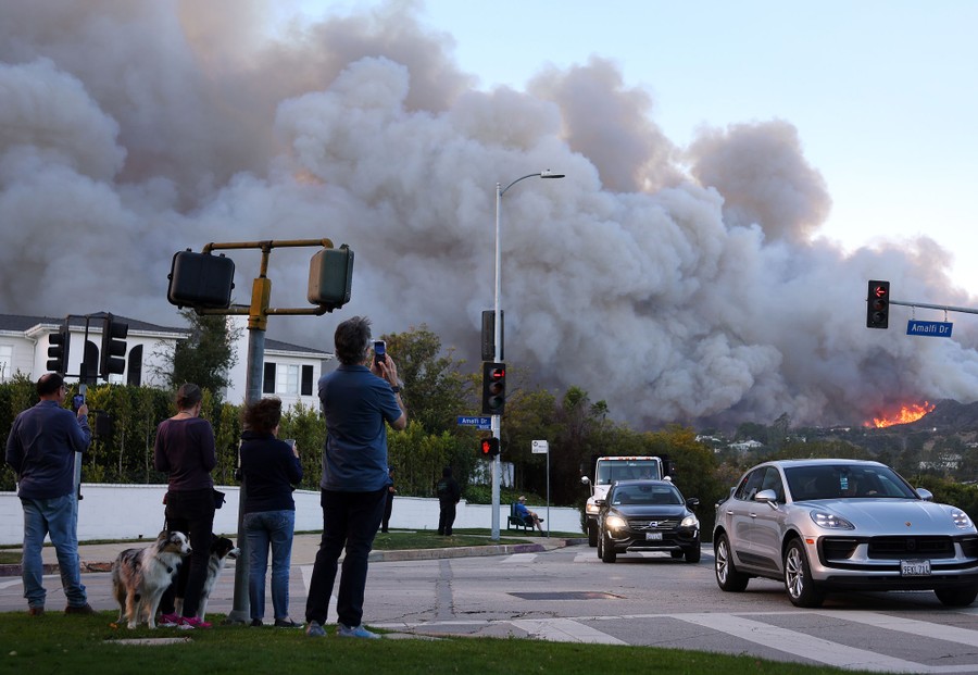 Several people stand on a street corner, watching smoke rise from a nearby wildfire.