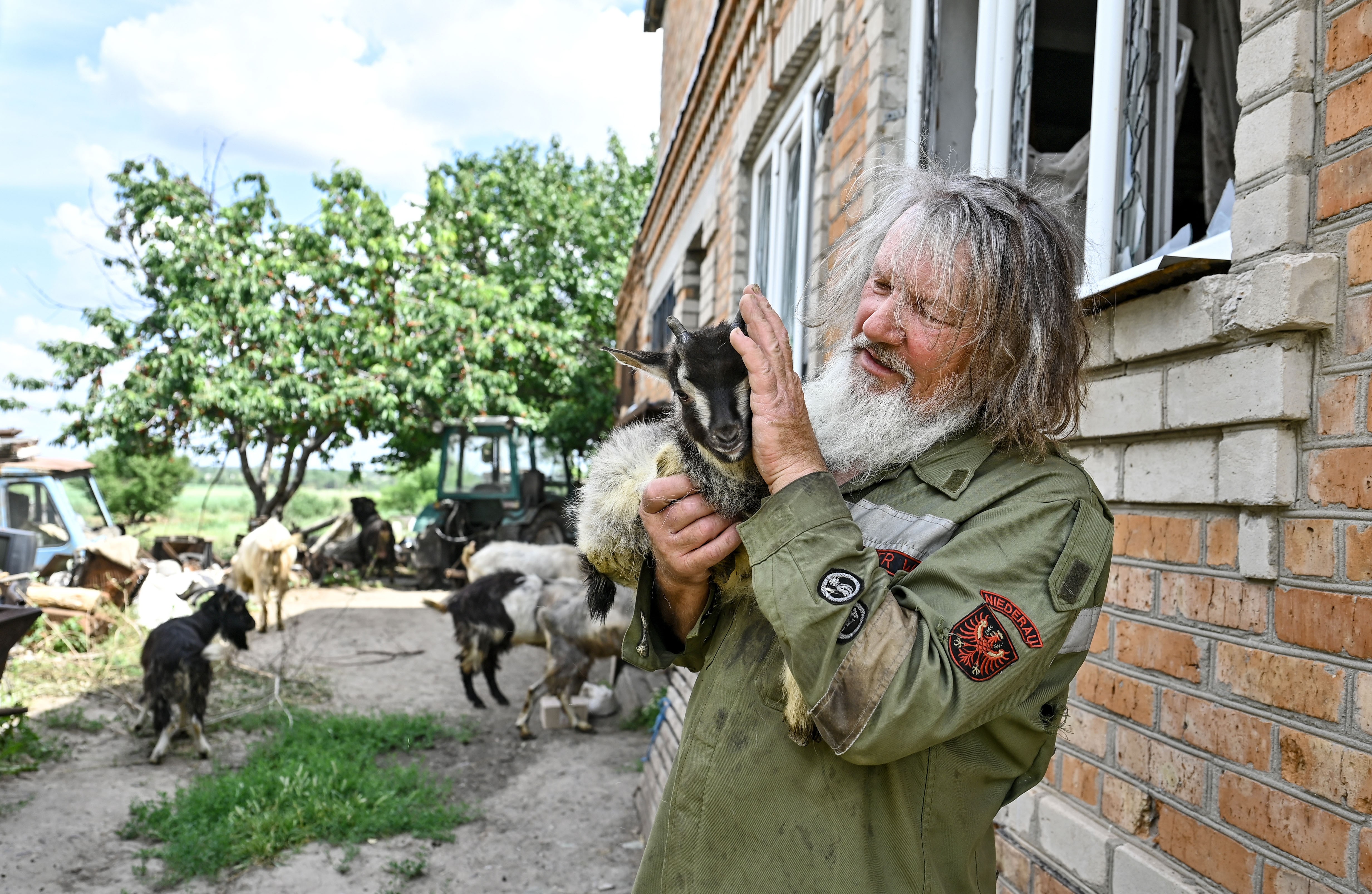 A man holds a young goat, standing near other goats beside a brick building.