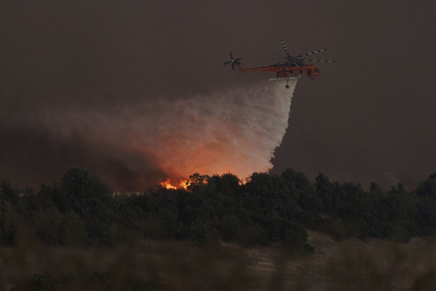 A helicopter drops water over a wildfire, the glow of the fire illuminating the water as it falls.