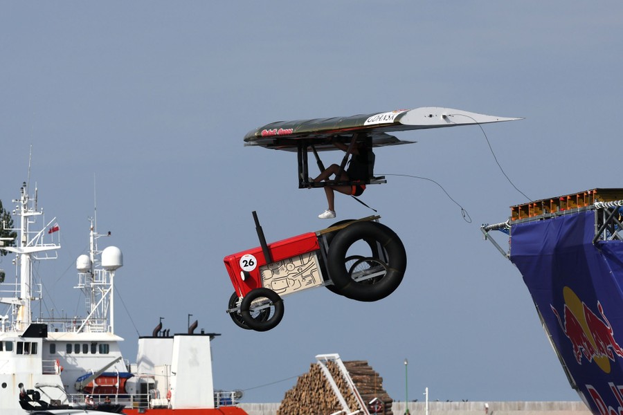 A person holds on to a hang glider while jumping off a ramp, as a hand-made tractor replica drops away beneath them.