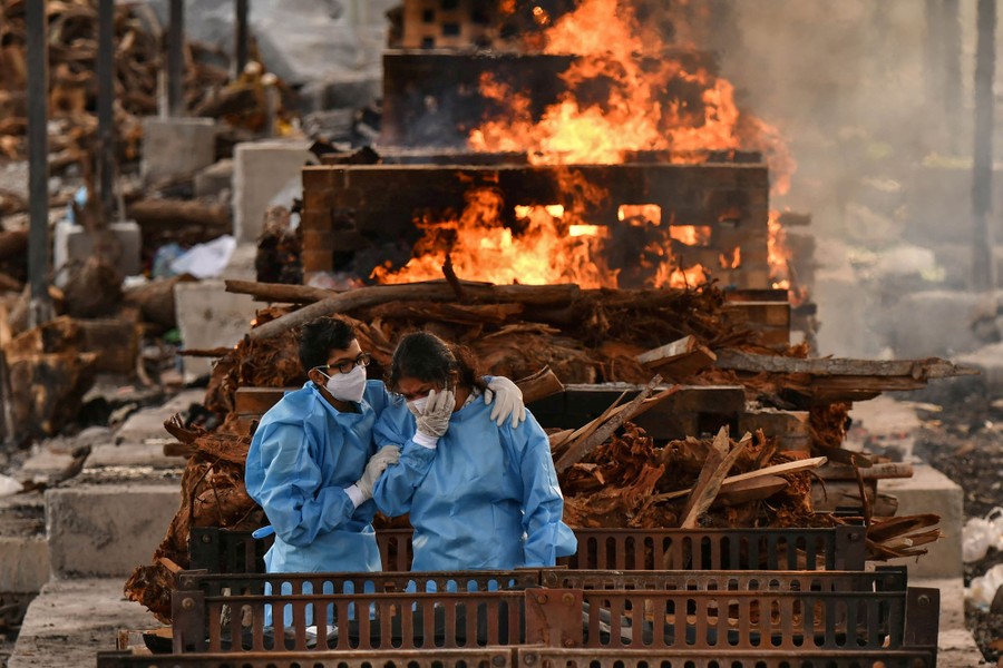 Two people wearing protective gear hold each other among several burning funeral pyres.