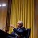 RFK Jr. expounds into a microphone while seated, against a backdrop of wood paneling and golden curtains