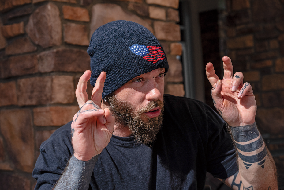 photo of bearded man in black beanie and black shirt talking and making an "air quote" gesture with heavily tattooed hands