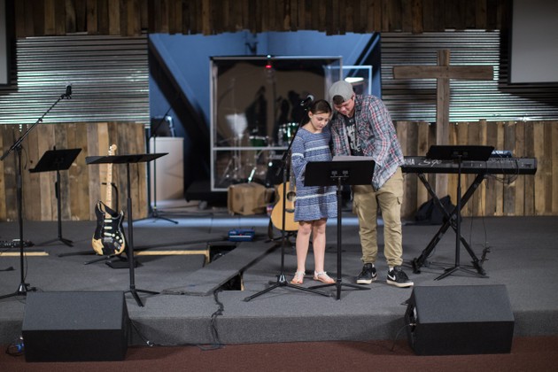 An older boy leans over his sister and a music stand. A cross is in the background alongside guitars and a keyboard.
