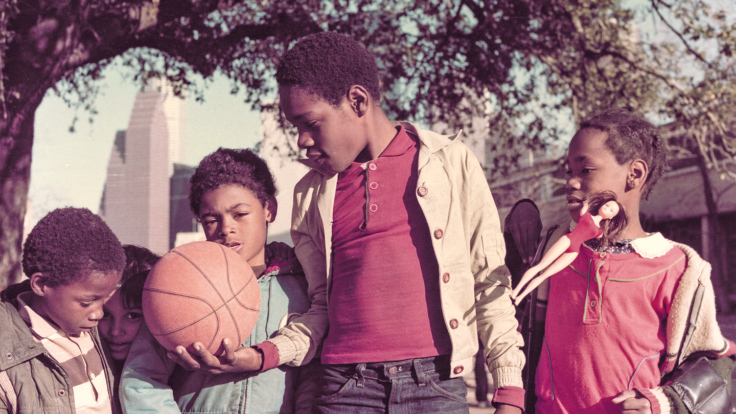 color photo of 5 children, one holding out basketball and another a Barbie doll, under large tree with Houston skyline in background