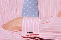 Close-up of a torso of a man wearing a pink dress shirt with cuff links and a blue tie, his arms crossed.