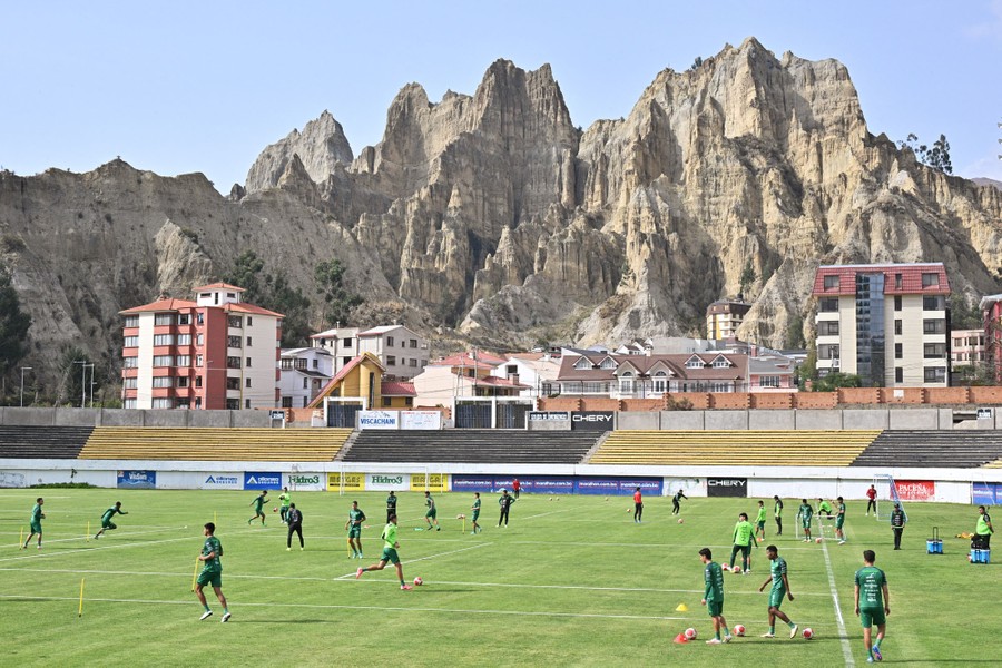 Soccer players practice on a pitch, backdropped by buildings and dramatic stone hillsides.