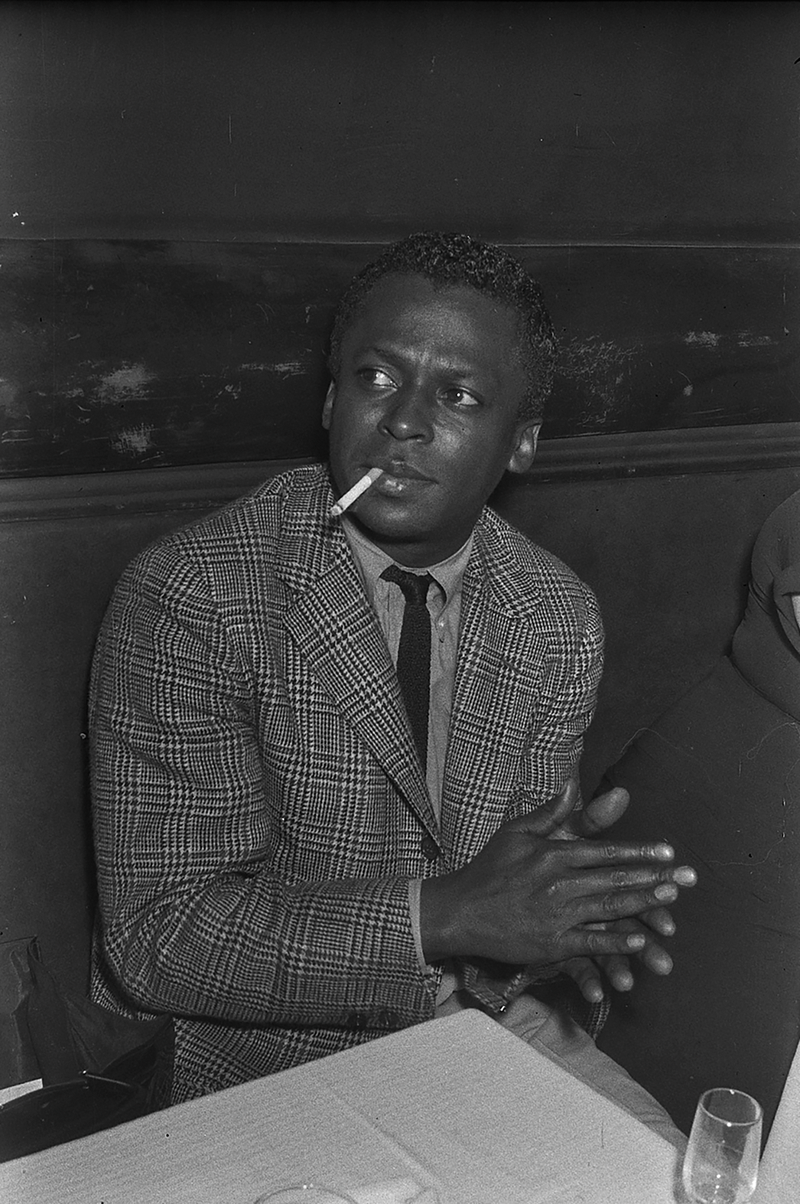 black-and-white photo of man in jacket and tie, sitting at table with cigarette in mouth and clapping hands