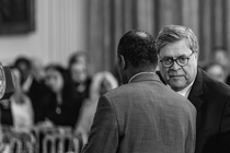 black-and-white photo of Bill Barr and Ben Carson conferring with each other with audience in background