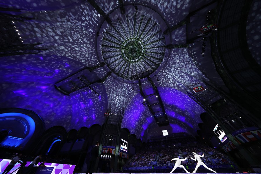 A wide view of a historical exhibition hall, its high ceiling illuminated by a lighting display, as a fencing match takes place before an audience.