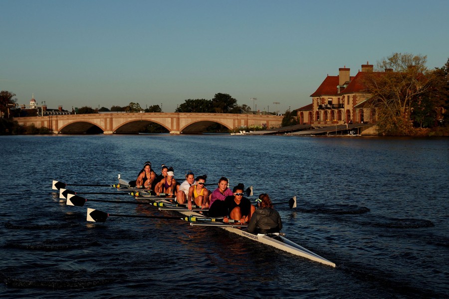 A crew team rows on the Charles River near a bridge and boathouse.
