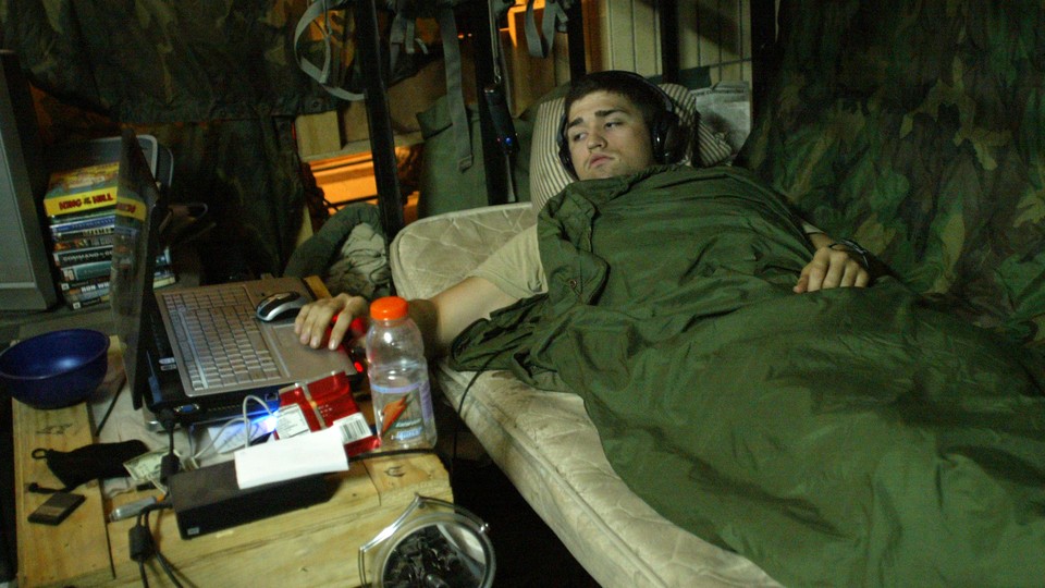 A man watches a movie on his laptop while wearing headphones and lying on a bed with camouflage bedding.