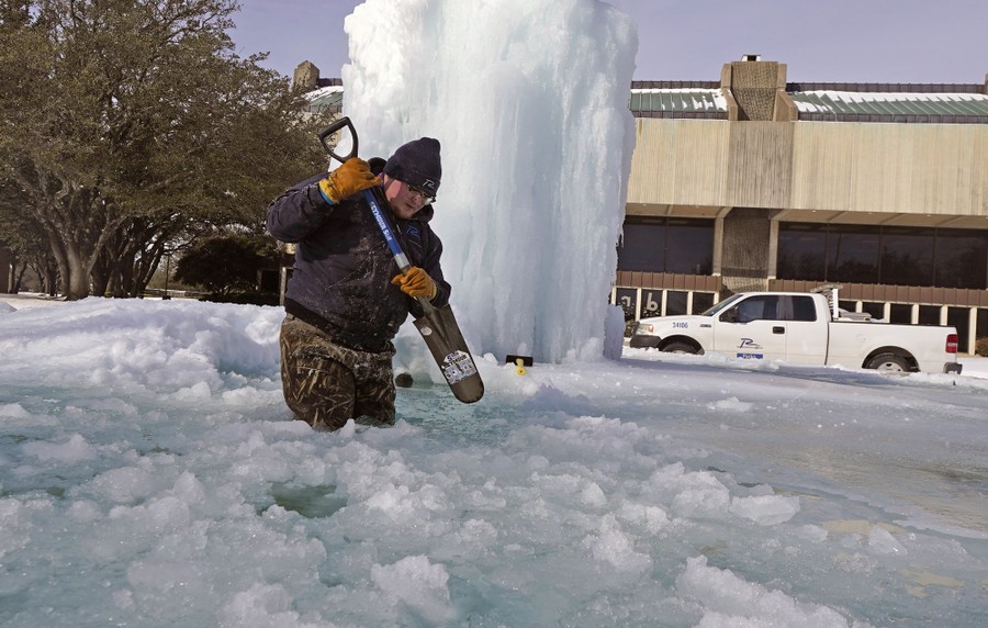 A person stands knee-deep in slushy water as they chop at ice in a frozen fountain.