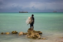 Kaitara Kautu, a net fisherman whose home flooded during last year's king tide in Betio, a town on the island of South Tarawa in Kiribati, stands on a rock in the middle of turquoise-colored water; a net rests on his left shoulder, and large ships sit in the distance.