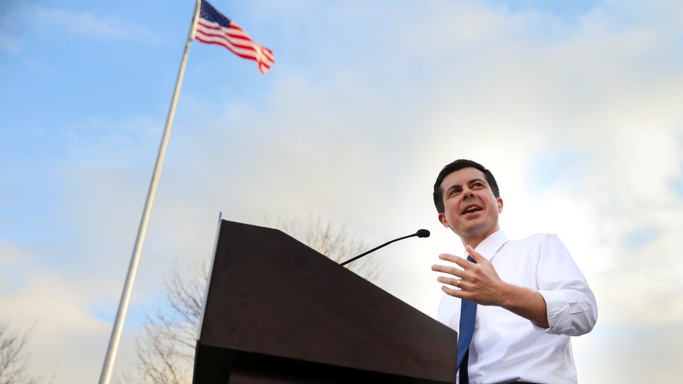 Pete Buttigieg speaks in Iowa with an American flag waving behind him and his wedding ring showing on his left hand