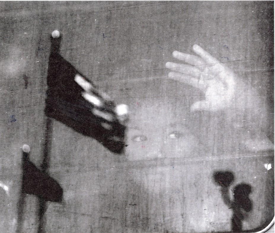 black and white photo of a young girl looking out of and pressing her hand against a window with the reflection of two American flags