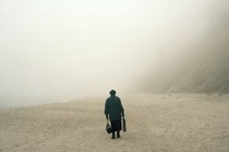 Color photograph of the back of a person in black clothing, carrying bags and standing on a beach