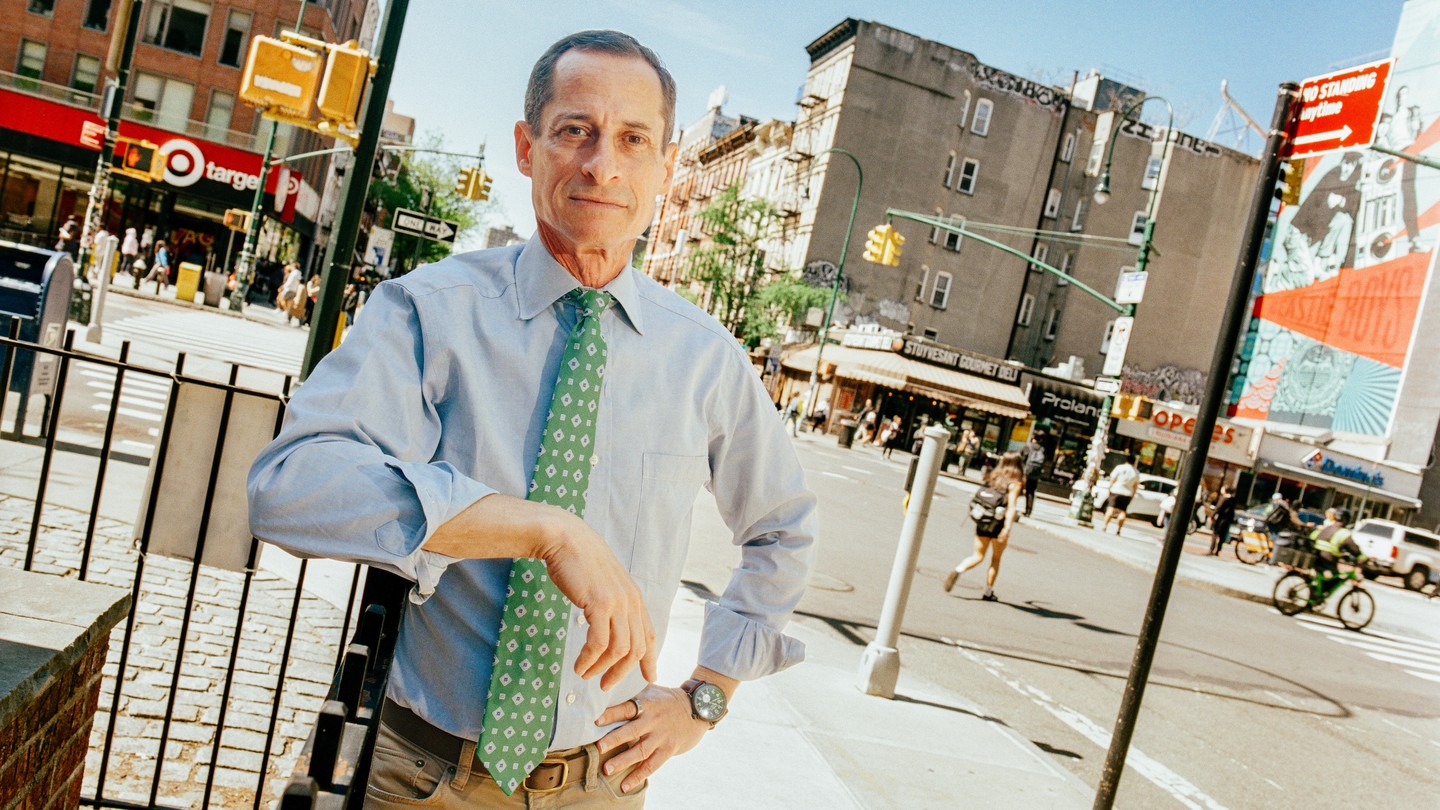 Color photo of Anthony Weiner standing near a busy urban intersection, his elbow resting on a fence.