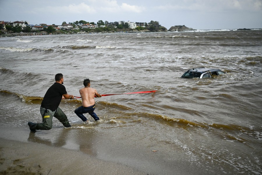 Two people pull on a strap that is attached to a mostly submerged car stuck in the surf.