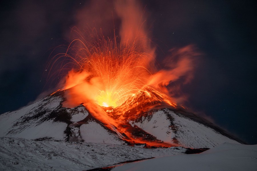 Lava erupts from the top of a snow-covered volcano, seen at night.