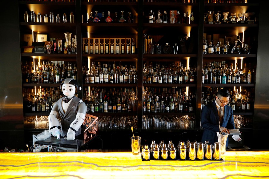 A man and a robot stand behind a well-stocked bar.
