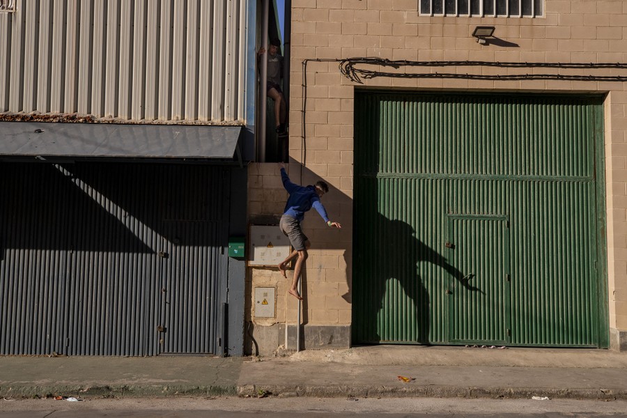 A migrant drops to the sidewalk from a narrow passage between buildings.