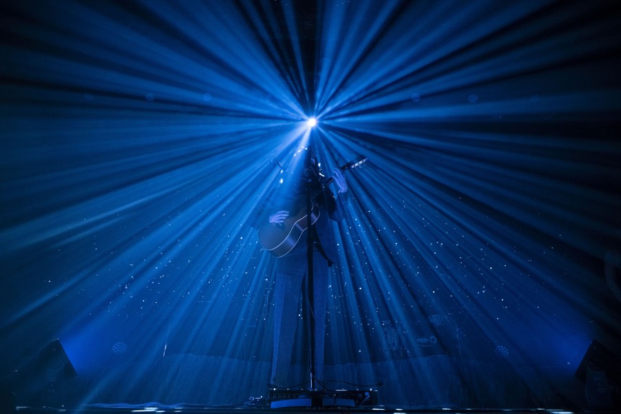 A performer plays a guitar onstage, backdropped by shafts of light shining from behind