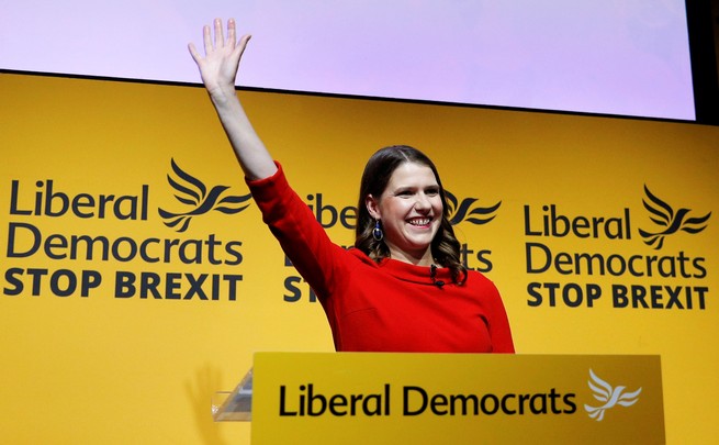 Jo Swinson raises her right hand behind a lectern and in front of signs for her Liberal Democrat party.