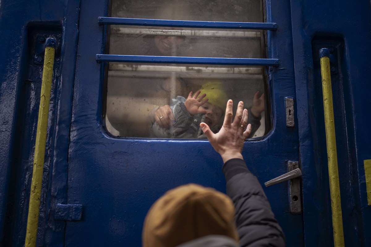 A father stands outside a train, reaching his hand up to his young son and wife, seen through a train window.