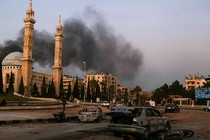 Photo of street view in Aleppo with smoke billowing above buildings and wrecked bombed cars in the foreground.