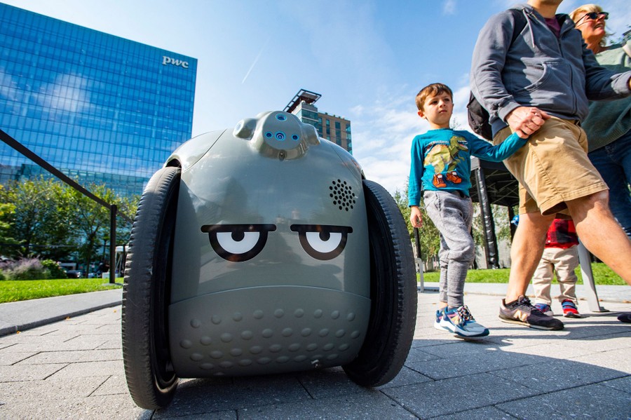 A grumpy-looking robot (angry eyes painted on its front) rolls beside several people.