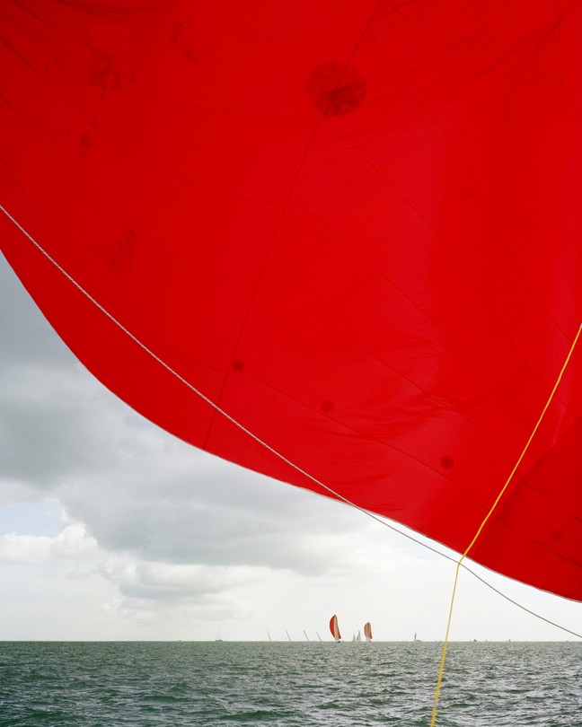 Boats with bright red sails on the ocean, one sail close up to the camera and covering the top half of the frame